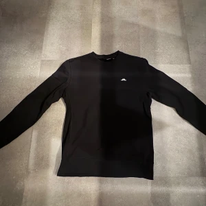 Svart tröja från J.Lindeberg - Säljer en stilren svart tröja från J.Lindeberg med en liten logotyp på bröstet. Tröjan har långa ärmar och en klassisk rund halsringning. Perfekt för en minimalistisk look. Aldrig använd och i riktigt bra skick.