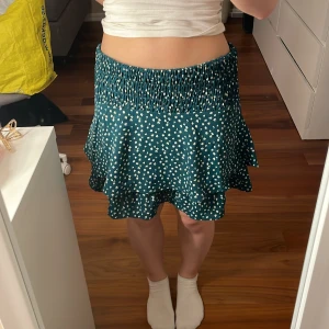 Grön blommig kjol från Grunt - Säljer en söt grön kjol från Grunt med vita blommor. Kjolen har en volangdetalj och en elastisk midja för en bekväm passform. Aldrig använt. Det står att storleken är XL men passar dig som är brukar ha xs/s