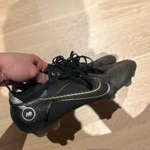 Svarta fotbollsskor från Nike - Säljer ett par svarta fotbollsskor från Nike med snörning och en stilren design. Skorna har en slitstark sula och är perfekta för fotbollsplanen. De har en diskret gulddetalj som ger en extra touch.