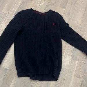 Svart stickad tröja från Ralph Lauren - Säljer en stilren svart stickad tröja från Ralph Lauren med det klassiska röda logotypbroderiet på bröstet. Tröjan har en rund halsringning och långa ärmar, perfekt för kyligare dagar. Passar både till jeans och chinos för en avslappnad look.