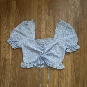 Blommig blus med volangdetaljer - Blommig blus i ljusblått med vita små blommor. Blusen har korta puffärmar och en knytning framtill som ger en fin rynkeffekt. Volangdetaljer vid ärmslut och nederkant. Toppen funkar lika bra som storlek S.