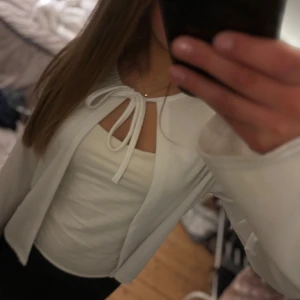 Vit bolerotopp med knytband - Säljer en stilren vit bolerotopp med knytband framtill. Perfekt att bära över en topp för en extra touch. Den har långa ärmar och är tillverkad i ett mjukt material.