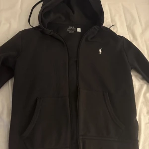 Svart hoodie från Ralph Lauren - Säljer en stilren svart hoodie från Ralph Lauren med dragkedja och huva. Den har en liten broderad logga på bröstet och praktiska fickor framtill. Perfekt för en avslappnad look.