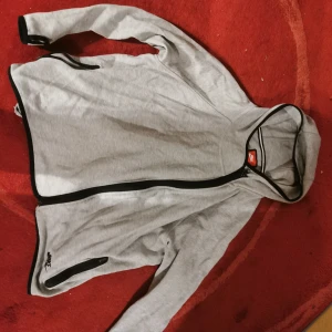 Grå vindjacka från Nike - Säljer en grå vindjacka från Nike med svarta detaljer. Jackan har dragkedja och långa ärmar. Perfekt för en sportig look eller en avslappnad dag ute.
