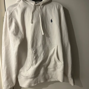 Vit hoodie från Ralph Lauren - Säljer en stilren vit hoodie från Ralph Lauren med det klassiska logotypbroderiet på bröstet. Tröjan har en känguruficka och justerbar huva med dragsko. Perfekt för en avslappnad look.