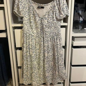 Blommig klänning från BikBok - Säljer en söt vit klänning med blått blommönster från BikBok. Den har korta ärmar och en v-ringning med knytband. Perfekt för en somrig look! Säljer den för att den inte kom till användning. 