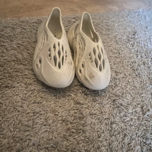 Beiga yeezy foam runners - Säljer ett par beige sandaler med unikt hålmönster. De är tillverkade i syntetmaterial och har en modern design som passar perfekt för varma dagar.