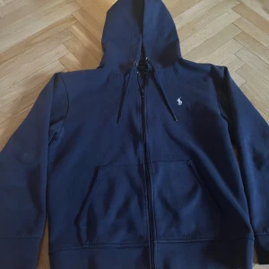 Marinblå hoodie från Ralph Lauren - Säljer en snygg marinblå hoodie från Ralph Lauren med dragkedja och broderad logga på bröstet. Den har en justerbar huva och praktiska fickor framtill. Perfekt för en avslappnad stil. tröjan är i storlek medium(175/84A) nypris är 2200. säljer för 500  priset kan diskuteras.