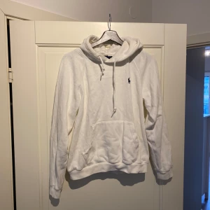 Vit hoodie från Ralph Lauren - Säljer en klassisk vit hoodie från Ralph Lauren med en liten blå logga på bröstet. Hoodien har en känguruficka och justerbar dragsko i huvan. Perfekt för en avslappnad stil.