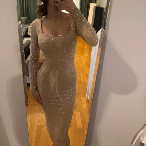 Champagnefärgad klänning med paljetter - Helt oanvänd med lapp kvar!! En elegant champagnefärgad klänning med glittrande paljetter. Den har en tight passform och långa ärmar, perfekt för en festlig kväll. Klänningen är dekorerad med små fyrkantiga och runda paljetter som ger en lyxig känsla.