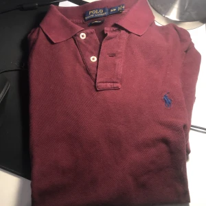 Vinröd pikétröja från Ralph Lauren - Säljer en stilren vinröd pikétröja från Ralph Lauren i storlek S. Tröjan har en klassisk krage med knappar och det ikoniska broderade logotypen i blått på bröstet. Perfekt för en avslappnad men ändå elegant look. Jätte jätte lite uttvättad, 6/10 skick