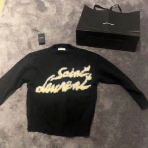 Svart tröja från Saint Laurent - Säljer en svart tröja från Saint Laurent med vit text på framsidan. Perfekt för en stilren outfit! Riktigt skönt stickat material bekvämt och varmt! Har av er vid frågor eller funderingar!