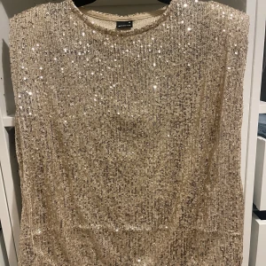 Glittrig topp från Gina Tricot - Snygg glittrig topp i champagnefärg från Gina Tricot. Perfekt för att glänsa på festen med sitt paljettöverdrag. Toppen har en rak passform och är ärmlös, vilket ger en elegant look. Ett måste för garderoben om du älskar att sticka ut! ✨