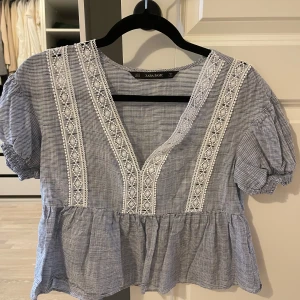 Blus från Zara Basic - Säljer en söt blus från Zara Basic i blått och vitt. Blusen har korta ärmar och v-ringning med vackra spetsdetaljer framtill. Perfekt för en avslappnad och stilren look.