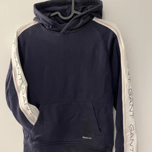 Mörkblå hoodie från Gant - Snygg mörkblå hoodie från Gant med vita och rosa detaljer längs ärmarna. Tröjan har en klassisk känguruficka och en bekväm huva. Inga skador eller fläckar inte alls mycket använd. 