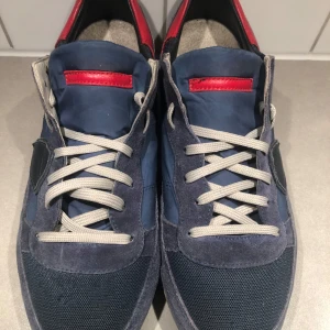 Philippe model TRPX - Detta är ett par Philippe Model TRPX-sneakers i marinblått med röda detaljer, vita skosnören och en mix av mocka, nylon och läder. De har en retroinspirerad design och visar visst slitage på sulorna. Bra skick bara något litet på sulan som inte syns. Storlek 44. Pris kan diskuteras vid en snabb affär 