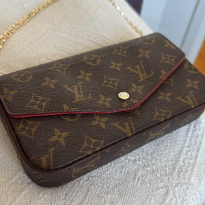 Brun monogram kuvertväska med kedja - Snygg brun kuvertväska med multifunktionellt guldfärgat axelband (kedja) med klassiskt monogrammönster. Väskan har en elegant knappstängning och röda detaljer längs kanterna. Väldigt bra kvalitet och perfekt för att ge en lyxig touch till din outfit.