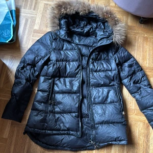 Svart tjock dunjacka med pälskrage - Säljer en svart tjock dunjacka med pälskrage och dragkedja. Jackan har en quiltad design och långa ärmar. Perfekt för kalla dagar med sin varma och skyddande stil. Går till rumpan o liiite till. Knappt använd så i väldigt bra skick. Känns även som luvan har ett exklusivt  o mjuk päls