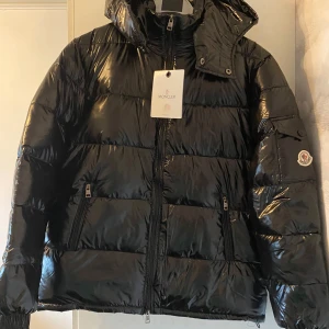 Svart pufferjacka från Moncler - Säljer en snygg svart pufferjacka från Moncler. Jackan har en glansig yta och är utrustad med dragkedja framtill. Den har en hög krage och en avtagbar huva. Perfekt för kyliga dagar och ger en stilren look. Kan gå ner i priset vid snabb affär. Skicka bud!