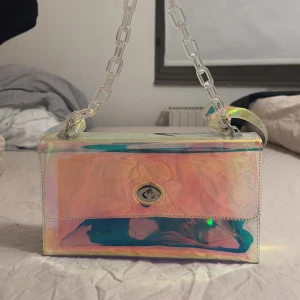 Iridescent axelväska med kedja - Snygg iridescent axelväska med en transparent kedja som axelrem. Väskan har en unik färgskiftande yta som fångar ljuset i olika nyanser. Den har ett vridlås framtill för säker förslutning. Perfekt för att ge din outfit en futuristisk touch.