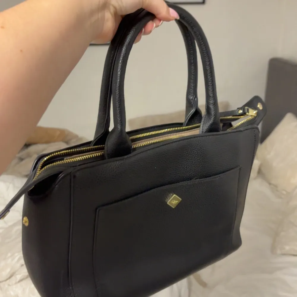 Snygg svart handväska i skinn med praktisk dragkedja och dubbla handtag. Väskan har en stilren design med en liten dekorativ detalj på framsidan. Perfekt för att bära dina dagliga nödvändigheter med stil.. Laukut & Käsilaukut.