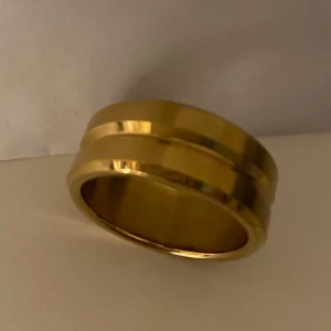 Fin Guldfärgad ring - Elegant guldfärgad ring med en stilren design. Ringen har en slät yta med två subtila linjer som löper runt omkretsen, vilket ger den en modern touch. Perfekt för att lägga till en touch av lyx till din stil. Aldrig använd 
