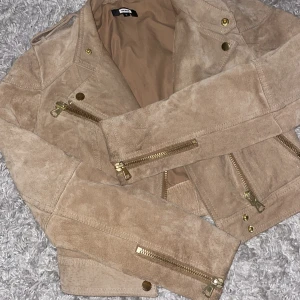 Beige mockajacka med dragkedjor - Snygg beige mockajacka från Bik Bok med guldfärgade dragkedjor och knappar. Jackan är perfekt för höst och vår. Den har en croppad passform och långa ärmar.