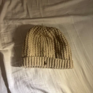 Beige stickad mössa från Ralph Lauren - Snygg beige stickad mössa från Ralph Lauren. Tillverkad i en mjuk ullblandning med ribbad kant. Perfekt för kyliga dagar. 80% wool 20% nylon. Skick 8,5/10