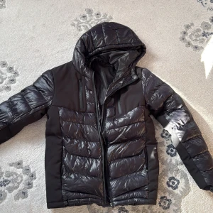 Svart pufferjacka från Brave Soul - Snygg svart pufferjacka från Brave Soul med huva och dragkedja. Jackan har en quiltad design och är perfekt för kyligare väder. Den är tillverkad i syntetmaterial och har en modern look.