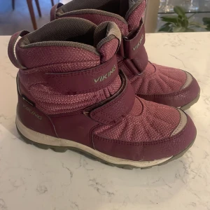 Rosa vinterstövlar från Viking - Säljer ett par mörk rosa vinterstövlar från Viking med praktiska kardborreband. Stövlarna har en robust sula för bra grepp och är perfekta för kalla dagar. De är designade för att hålla fötterna varma och torra under vintern.