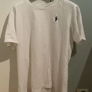 Ralph Lauren Tröja T-shirt - Säljer en klassisk vit t-shirt från Ralph Lauren med den ikoniska broderade loggan i mörkblått på bröstet. Perfekt för en stilren och avslappnad look.