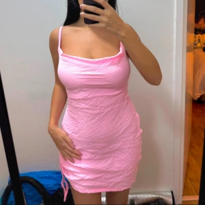Rosa kortklänning med öppen rygg - Säljer en söt rosa kortklänning med smala axelband och en stilren öppen rygg. Klänningen har en åtsittande passform och är perfekt för sommarens varma dagar. Den har en lätt rynkad textur som ger en extra touch av stil.