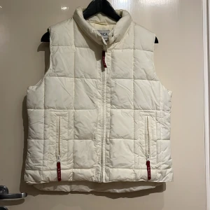 Vit quiltad dunväst från Amica - Säljer en stilren vit quiltad dunväst från Amica. Västen har en dragkedja framtill och praktiska fickor med röda detaljer. Perfekt för kyligare dagar när du vill hålla värmen utan att kompromissa med stilen.
