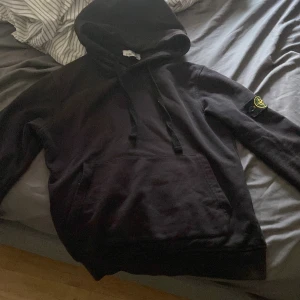 Svart hoodie från Stone Island - Säljer en stilren svart hoodie från Stone Island med deras ikoniska märke på ärmen. Tröjan har en klassisk design med huva och känguruficka framtill. Perfekt för en avslappnad look.