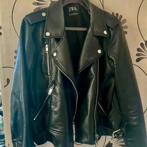 Ny Svart skinnjacka från Zara - Snygg svart skinnjacka från Zara med dragkedjor och knappar. Jackan har en klassisk bikerstil med bälte i midjan och axelklaffar. Perfekt för en cool look!