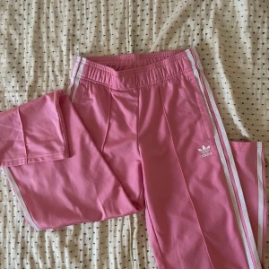 Rosa träningsbyxor från Adidas - Snygga rosa träningsbyxor från Adidas med vita ränder längs sidorna. Byxorna har en elastisk midja med snörning för extra komfort. Kan ej köpas i butik eller på nätet. Sitter Low waist. Barnstorlek men sitter som XS!
