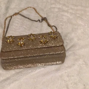 Glittrig guld axelväska med blommor - Snygg glittrig guld axelväska med dekorativa blommor på framsidan. Väskan har en elegant kedjerem och är perfekt för att ge en touch av glamour till din outfit.