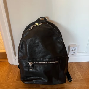 Svart ryggsäck med dragkedja från Zara - Säljer en svart ryggsäck från Zara i syntetmaterial med silverfärgade dragkedjor. Ryggsäcken har justerbara axelremmar och en praktisk framficka med dragkedja. Perfekt för vardagsbruk och enkel att matcha med olika stilar.