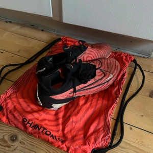 Röda fotbollsskor från Nike - Snygga röda Nike Phantom fotbollsskor med svart snörning och mönster. Perfekta för fotbollsplanen med en stilren design och bra passform.
