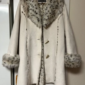 Vit kappa med fuskpäls från KRISS Sweden - Elegant vit kappa från Kriss Sweden med mjuk fuskpälsdetalj på krage och ärmslut. Kappan har en klassisk design med knappar framtill och är perfekt för dig som letar efter en snygg kappa. Jackan är i storlek 42 men sitter bra och passar mig som är en 36/38. 