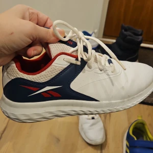 Vita sneakers från Reebok - Säljer ett par vita Reebok sneakers med blå och röda detaljer. Skorna har snörning och en bekväm sula, perfekt för vardagsbruk. De är stilrena och lätta att matcha med olika outfits.