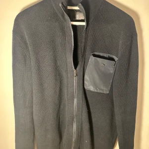 Svart stickad kofta med dragkedja - Snygg svart stickad kofta från Less Worries med dragkedja och en praktisk bröstficka med knapp. Perfekt för kyliga dagar och ger en stilren look. Tyvärr är knappen på bröstfickan sönder