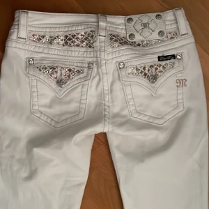 Vita jeans med paljettdetaljer från Miss Me - Snygga vita jeans från Miss Me med glittrande paljettdetaljer på bakfickorna och bältet. Byxorna har en lågmidjad passform och är dekorerade med knappar och broderade detaljer. Perfekta för att sticka ut med en unik stil!!💓💓Midjemått rakt över: 37cm, innerbenslängd, 75cm. De är skinny men är enkla att sy ut till bootcut. Säljer bara då färgen inte passar mig!