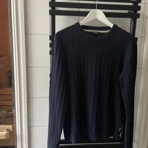Mörkblå stickad tröja från Jack & Jones - Säljer en stilren mörkblå stickad tröja från Jack & Jones. Tröjan har ett klassiskt kabelstickat mönster och långa ärmar med ribbade muddar. Perfekt för kyligare dagar. Nypris 500kr, säljer för 249kr. Kan gå ner i pris vid snabb affär.