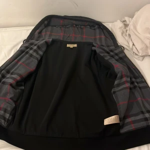 Svart hoodie från Burberry - Snygg svart hoodie från Burberry med dragkedja och klassiskt rutigt mönster på insidan av huvan. Perfekt för en stilren look med en touch av lyx. Den har praktiska fickor framtill och en justerbar huva med snören. Kan gå ner i pris 