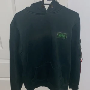 Mörkgrön hoodie från Alpha Industries - Snygg mörkgrön hoodie från Alpha Industries med stor vit logga på ryggen och bröstet. Tröjan har en praktisk ficka på ärmen och en justerbar huva utan snörning. Perfekt för en casual look.