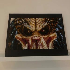 Skräcktavla, predator. Pärlplatta  - En pixelkonsttavla med ett motiv av ett ansikte i mörka och jordnära färger, inramad i svart. Tavlan har en unik och detaljerad design som ger ett kraftfullt intryck.                                    50 x 40 cm.