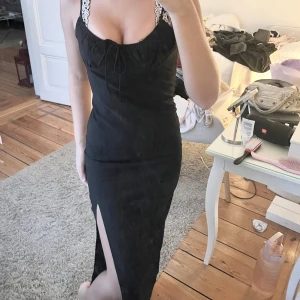 Svart klänning med spetsdetaljer - Elegant svart klänning med en djup slits framtill. Klänningen har en åtsittande passform och knytning vid bysten för extra stil.