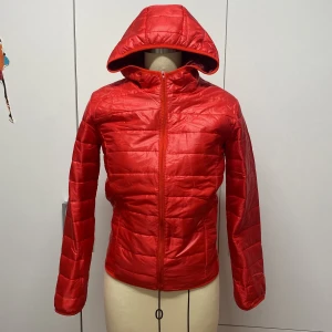 Röd quiltad pufferjacka med huva - Snygg röd pufferjacka med quiltat mönster och praktisk huva. Jackan har en dragkedja framtill och långa ärmar.