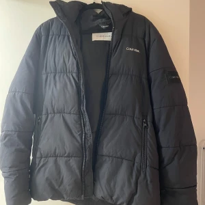 Svart pufferjacka från Calvin Klein - Snygg svart pufferjacka från Calvin Klein med dragkedja och broderad logga på bröstet och ärmen. Jackan har en hög krage och är perfekt för kyligare dagar. Använd 2 vintrar, kommer ej till användning längre.